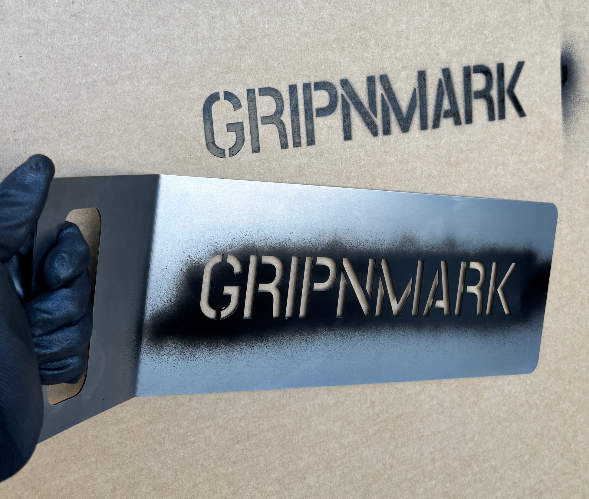 GRIPNMARK branding on a metal stencil held by a gloved hand against a cardboard background
