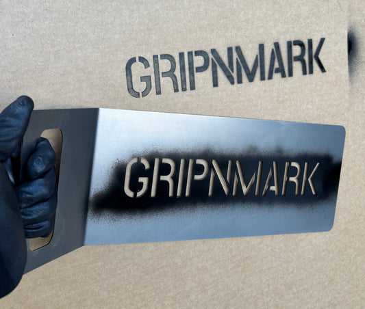 GRIPNMARK branding on a metal stencil held by a gloved hand against a cardboard background