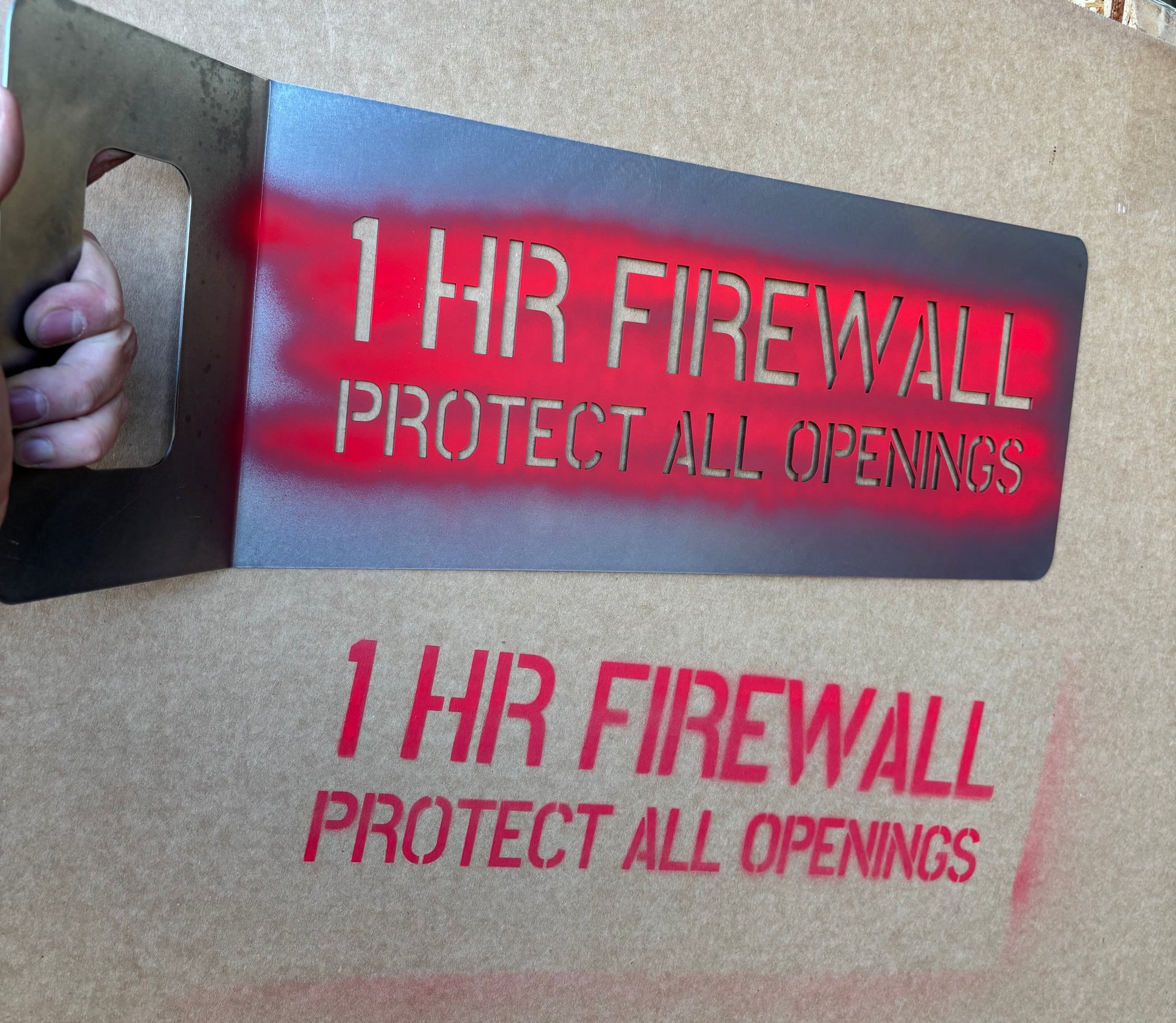 Reusable stencil with '1 HR FIREWALL PROTECT ALL OPENINGS' text painted on a cardboard background