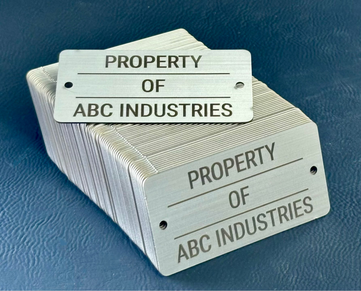 Stack of bulk matching metal tags with 'Property of ABC Industries' text on a blue background